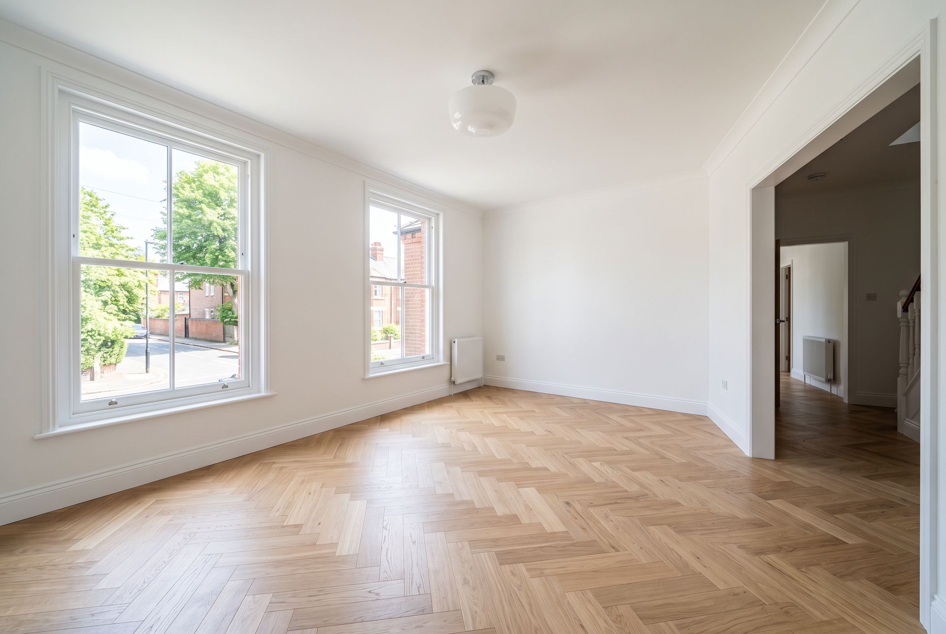 An empty living room with white walls, herringbone oak parquet and two sash windows