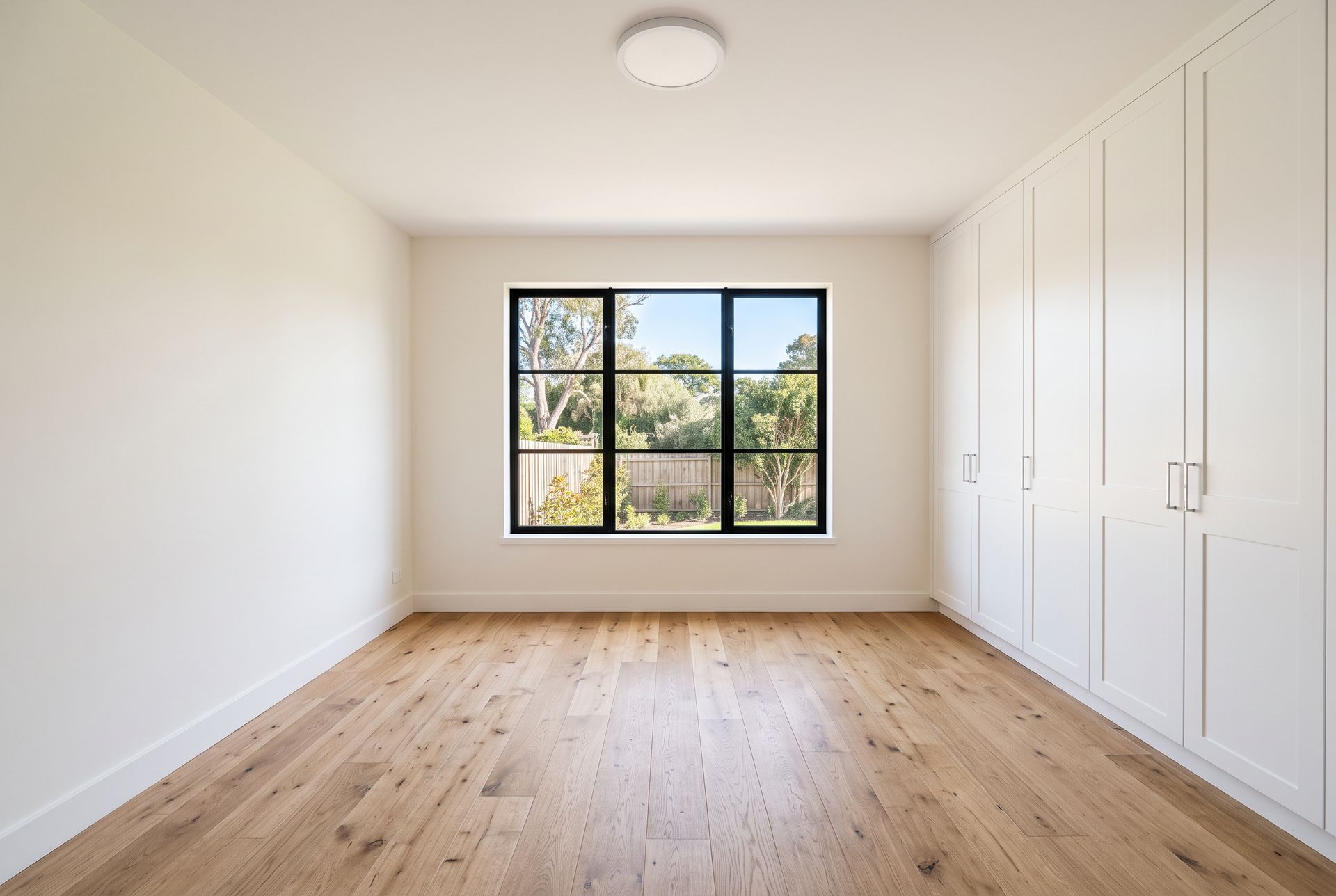 An empty bedroom with oak floors, a black framed window to the garden and a built-in white wardrobe